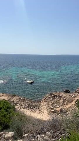 A view of the ocean from a cliff overlooking the water. The water is a deep blue with lighter turquoise patches near the shore. There are two small objects visible in the water, one appearing to be a floating debris or a small boat, and the other a darker object further offshore. The cliff face is rocky and covered with sparse vegetation. The sky is clear and blue.