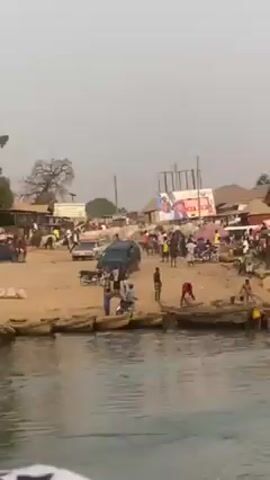 The video shows a riverbank community with a mix of people, boats, and vehicles. The camera initially focuses on the bustling scene on the sandy bank.  There are people of all ages, some standing by the water, others walking around. Several small, simple boats are docked along the shoreline.  A dark SUV and a few motorbikes are also visible in the sandy area, suggesting vehicular traffic. A large billboard with faces is visible in the background.  The camera then shifts to show a person in a blue shirt and backpack, looking towards the riverbank. The opposite bank shows more of the same kind of environment with trees, buildings, and people gathered around. The overall atmosphere is active and communal, typical of a riverside village.