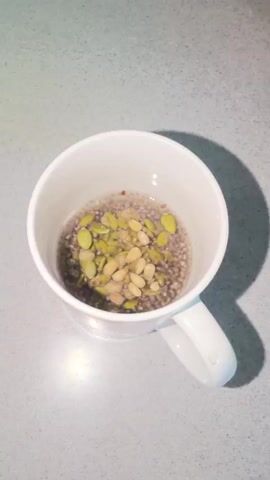 A white ceramic mug sits on a gray speckled countertop. Inside the mug, there is a mixture of chia seeds and what appear to be pumpkin seeds and possibly other larger seeds, suspended in a clear liquid. An immersion blender is being inserted into the mug. The blender's metal shaft is submerged into the mixture. Water is being poured from a black appliance, identified by a label with Cyrillic text and "CE" certification, into the mug, covering the seeds and the blender head. The blender is then activated, and the mixture is agitated and appears to be becoming a more uniform, reddish-brown color, likely due to the seeds breaking down.