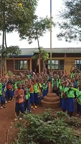 A group of young children in green shirts and blue skirts or shorts are gathered around a flagpole in front of a building. The children are clapping and raising their hands in the air, seemingly singing or chanting. A man in a similar green shirt stands slightly apart from the group, possibly leading them. The setting appears to be a schoolyard with trees and greenery visible. The building behind the children has several windows and a simple roof.