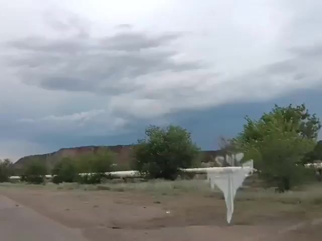 The video shows a landscape under a cloudy sky. In the foreground, a dirt road is visible on the left. A long, white pipe runs horizontally across the middle ground, supported by what appear to be concrete structures. Green bushes and dry grass are scattered along the pipe. In the background, a large, brown, barren hill or mountain is visible. The sky is filled with heavy, dark clouds, suggesting impending rain or stormy weather. There are no people or animals visible in the video. There is a distorted reflection on the lens of the camera.