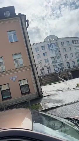 The video shows a street view of buildings. On the left is a pale pink building with several windows and a small balcony. A street sign with Cyrillic text and the number 42 is visible. To the right of this building is a more modern, light-colored building with a glass entrance and stairs leading up. Between the two buildings is a darker, older building. Russian flags are visible on the older building. In the foreground, the hood and windshield of a car are visible, suggesting the video is being filmed from inside a vehicle. The sky is cloudy. The street appears to be wet, possibly from recent rain. A white car is visible further down the street.