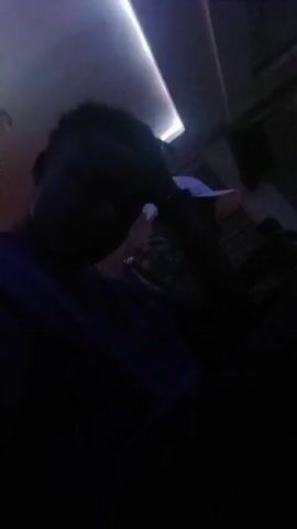 The video shows two dark-skinned individuals in what appears to be a dim room with colorful lighting, possibly a bar or lounge. The person in the foreground is wearing dark clothing, while the other person, slightly behind, wears a white baseball cap. The room has a modern design with linear ceiling lights that cycle through blue, purple, and white. The camera angle is from a low perspective, looking slightly up at the subjects. There is music playing in the background, suggesting a social atmosphere.