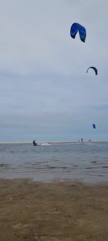 The video shows a wide shot of a beach and a body of water. In the foreground is a sandy beach. In the middle ground, on the water, a person is kitesurfing. The person is wearing dark clothing and is riding on a board, pulled by a kite. The kite is blue and appears to be in the sky above the kitesurfer. In the background, there is a long stretch of sand or land, with a few more people standing near the water's edge. The sky is overcast with clouds. There is another kite visible in the upper part of the frame, which is also blue. The water is choppy, with small waves. The overall scene depicts a recreational activity on a cloudy day at the beach.