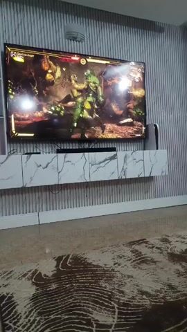 The video shows a TV screen displaying a Mortal Kombat match. The characters are fighting in a jungle-like environment. The text "FINISH HIM" is displayed in large red letters. The TV is mounted on a white marble-patterned stand against a gray striped wall. A PlayStation 5 console is visible to the right of the TV. The floor is covered with a glossy tile and a decorative rug. The camera is positioned low and close to the ground, providing a view of the room.