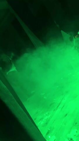 The video shows a room lit in green light with smoke. There are wooden boards visible on the floor. The ceiling appears slanted with exposed beams. There are no people visible. The source of the smoke and green light is not visible.