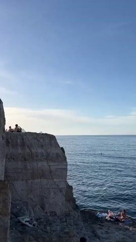 The video displays a rocky cliff next to a blue ocean under a clear sky with scattered clouds. Two individuals are seated on the cliff's edge, gazing out at the sea; one is a man wearing sunglasses, and the other is a woman. A white plastic chair is discarded on a lower part of the cliff. In the distance, a person is swimming in the ocean, and two other individuals are lying on towels on a lower rock platform near the water. Small landmasses are visible on the horizon.