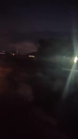 A dark, low-light video shows an outdoor scene. The camera appears to be moving, possibly in a vehicle. Several streetlights and car headlights are visible, creating lens flares and streaks of light. In the background, there are indistinct shapes that could be buildings or trees. The sky is dark, with some clouds faintly illuminated by the lights. The overall impression is that of a night-time drive through a suburban or rural area.