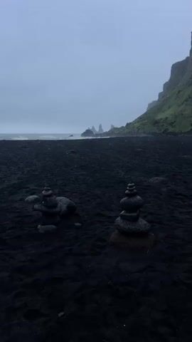 The video shows a black sand beach with two stacks of stones, a cliff face covered in green vegetation, and rock formations in the distance, under a cloudy sky. The beach is largely empty. The stacks of stones are in the foreground. The ocean is in the background.