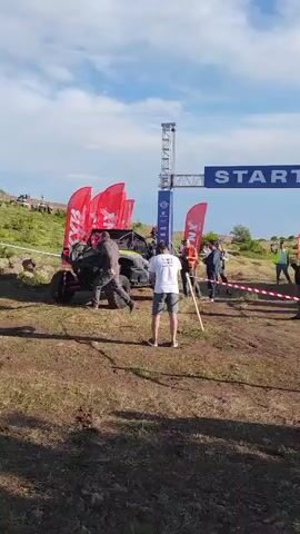 The video shows an off-road vehicle, likely a UTV or ATV, parked at a starting line. A large blue banner with the word "START" in white capital letters is suspended above the track, supported by a metal gantry. Several red flags with a white logo are planted behind the vehicle.

There are several people in the scene. A person in a white t-shirt and grey shorts stands facing away from the camera, holding a wooden pole. Another person in a grey jacket is walking past the vehicle on the left. Other individuals are visible standing or walking near the starting line and in the background.

The setting appears to be a dusty, outdoor course with some dry grass and uneven terrain. In the distance, there are rolling hills and a few structures. The sky is partly cloudy with blue patches. The lighting suggests it is daytime, likely late morning or early afternoon. The overall atmosphere seems to be that of a sporting event, possibly a race or competition, with participants and spectators gathered.

The visible text on the banner clearly reads "START." The logos on the red flags are not entirely clear but appear to be stylized. There is also some text on a banner attached to the gantry, but it is too small to read. The scene is set in Antarut, Armenia.