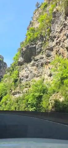 The video shows a car traveling on a road cut into a mountain pass on a sunny day. The road is paved and has a concrete barrier separating it from a drop-off. The mountains are tall and rocky, with some green vegetation growing on them. The sky is blue with a few white clouds. The camera is mounted inside the car, looking out the front windshield.