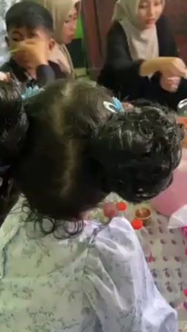 The video shows a group of people, including children and adults, sitting on the floor around a table in what appears to be a home. The children are eating, and some adults are holding plates and phones. There are bowls and containers of food on the table, as well as bottles of water. The room is decorated with pictures on the wall and has green paint. The people are dressed in a variety of clothing, including headscarves.
