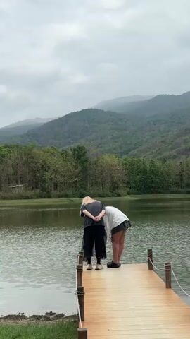 Two young women are standing on the end of a short wooden pier, looking down at a lake. The woman on the left has long, blonde hair and is wearing a dark t-shirt, black pants, and sneakers. Her hands are clasped behind her back. The woman on the right is wearing a white t-shirt, grey pleated skirt, and dark shoes, and is bent slightly forward, looking into the water. Behind them is a calm lake, surrounded by green trees. Mountains are visible in the misty background, and the sky is overcast. The scene is serene.