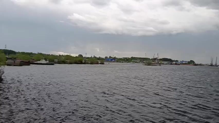 The video captures a wide expanse of dark, choppy water under an overcast sky. On the left, a distant shoreline is visible with a line of green trees, scattered buildings, and what appear to be several industrial cranes. In the foreground on the right, a large, weathered white and grey metallic structure, resembling a crane or a davit system for a lifeboat, dominates the frame. It features perforated metal grates, various cables, and a large, rusty spool or wheel. The camera appears to be on a moving vessel or platform, as there is a gentle swaying motion. The sky is heavily clouded, suggesting an overcast day. The sounds in the video include a distinct wind noise transitioning into a low rumble, possibly from an engine or machinery. The setting is a body of water in Igrim, Russia, with a distant industrial area along the shore. No discernible text is visible.