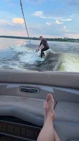 A man in a wetsuit and life vest is wake surfing behind a boat. He is riding on a light blue board and holding onto a rope attached to the boat. The boat is moving through water, creating a large wake. The camera is positioned inside the boat, looking out at the man. A person's legs are visible in the foreground, resting on the boat's seat. The sky is blue with some clouds, and there is a tree line in the distance. The boat has the brand name "CENTURION" visible on the seat.