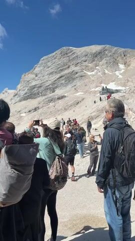 This video shows a group of people standing in a line, seemingly waiting for something, with a large mountain range in the background. The mountain slopes are covered in rock and some snow, and the sky is blue with scattered clouds. Several individuals are holding up their smartphones to take pictures or videos. The foreground features a woman with dark hair wearing a pink shirt and a grey backpack, also holding a phone. Behind her, a person in a green shirt and another in a backpack are visible. Further back, more people are gathered, and a band in traditional attire is playing music in front of the mountain. A tall pole with flags or banners stands to the left of the band. The ground is a rough, gravelly surface.