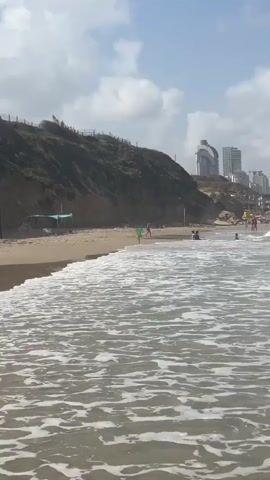 The video shows a beach with waves crashing on the shore. There are people on the beach and in the water, as well as some buildings in the background. The sky is blue with some clouds. The camera is positioned at the water's edge, looking out towards the ocean. The waves are small to medium in size and appear to be breaking on the sand. Some people are visible in the distance, enjoying the beach. There is a grassy cliff to the left with a few people and some structures, including a blue tarp. Further down the beach, buildings can be seen against the skyline. The water is clear and the sand is light brown. The overall atmosphere is one of a sunny day at the beach.