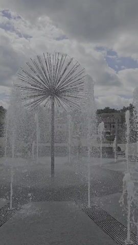 The media file shows a public space with a dandelion-shaped fountain. The fountain is the central object, with water spraying in a circular pattern around a metal structure that resembles dandelion seeds. In the background, there are buildings that appear to be in an urban setting, and trees. The sky is cloudy, suggesting it might be an overcast day. There are a few people visible in the distance, walking around the fountain. The ground is paved with a pattern of grey and black tiles, and there are some grate-like structures within the paving. The overall mood is peaceful and serene, with the sound of the water likely contributing to a calming atmosphere. No text is visible in the media file. The location is Bryansk, Russia.