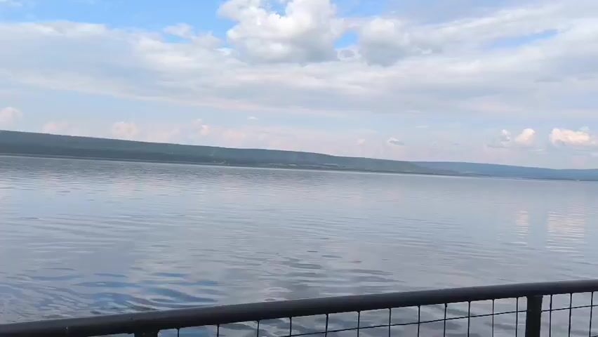 The video shows a wide expanse of calm water under a partly cloudy sky. The distant shore is visible as a low, dark line against the horizon. In the foreground, a black metal railing with a grid pattern is visible, suggesting the perspective is from a deck or balcony overlooking the water. The water is mostly still, with subtle ripples reflecting the sky.