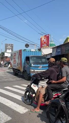 The video shows a street scene on a sunny day. In the foreground, a motorcycle with two occupants is stopped at a crosswalk. The driver, wearing a dark hoodie and a tan baseball cap, has his left arm extended as if signaling. The passenger, wearing a gray t-shirt and a green baseball cap, is looking to the right. Behind the motorcycle, a large truck with a white and red advertisement for "Cleo" is stopped. Further down the street, other vehicles, including cars and yellow vans, are visible. Overhead, power lines crisscross the sky, and various billboards and signs are mounted on buildings. The overall impression is a busy urban intersection.