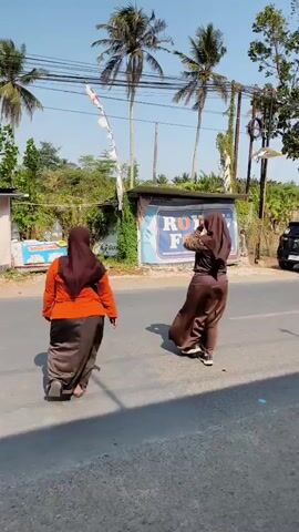 Two people wearing hijabs walk along a road. The person in front wears an orange top and a brown skirt. The person behind wears a brown outfit. A black car is parked on the side of the road, and a traffic cone is placed next to it. There are trees and buildings visible in the background, and the sky is clear and blue.