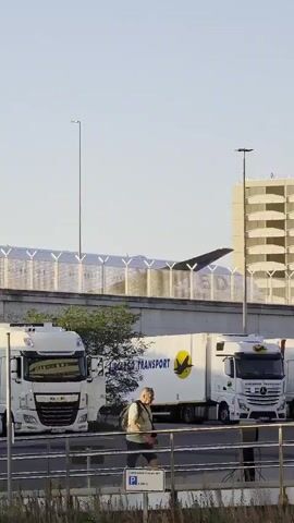 The video shows a man walking along a railing in front of a row of parked semi-trucks. The trucks have various company logos, including "Lufthansa Cargo" and "DAF." In the background, there is a large building with a repeating architectural pattern and a portion of an airplane wing with a blue and white logo visible above a fence. The scene appears to be an outdoor area, possibly a loading dock or parking lot, with streetlights and a clear sky. The man is wearing a light-colored shirt, shorts, and a backpack. He is walking from left to right across the frame.