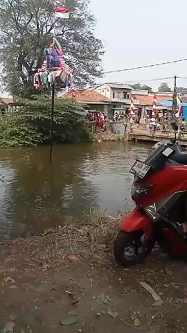 A red scooter is parked on the side of a muddy path. In the background, a body of murky water is visible. On the far side of the water, there is a bridge with several people on it, as well as buildings. A tall pole stands in the water, adorned with Indonesian flags and colorful decorations. More people are gathered on the bank of the river. The overall atmosphere appears festive.
