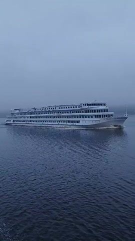 A long white river cruise ship is underway on a body of water under an overcast sky. The ship has multiple decks and many windows. The water is slightly choppy. The ship leaves a small wake behind it.