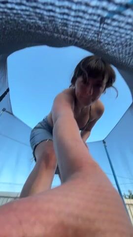 A person in a bikini top and denim shorts is on a trampoline, looking up into the sky. The camera is positioned below the person, looking up through the netting of the trampoline enclosure. The person jumps, and the camera moves rapidly, making the image blurry. The sky is blue with some clouds. The scene appears to be outdoors during the daytime.