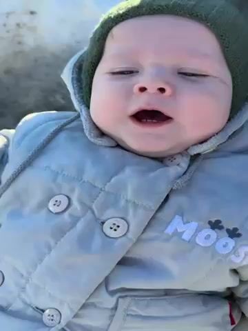 A baby wearing a gray snowsuit and green knit hat is seated in a stroller. The baby is vocalizing with an open mouth and appears to be making sounds or attempting to speak. The background shows a snowy or icy surface. The snowsuit has the word "MOOSE" embroidered on it, along with small floral designs.