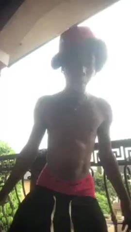 A young, dark-skinned man stands on a balcony with ornate black railing, shirtless and wearing black sweatpants and a red cap. He smiles and dances to music, sometimes looking at the camera. The sky behind him is bright with clouds. In the background, trees and buildings are visible.