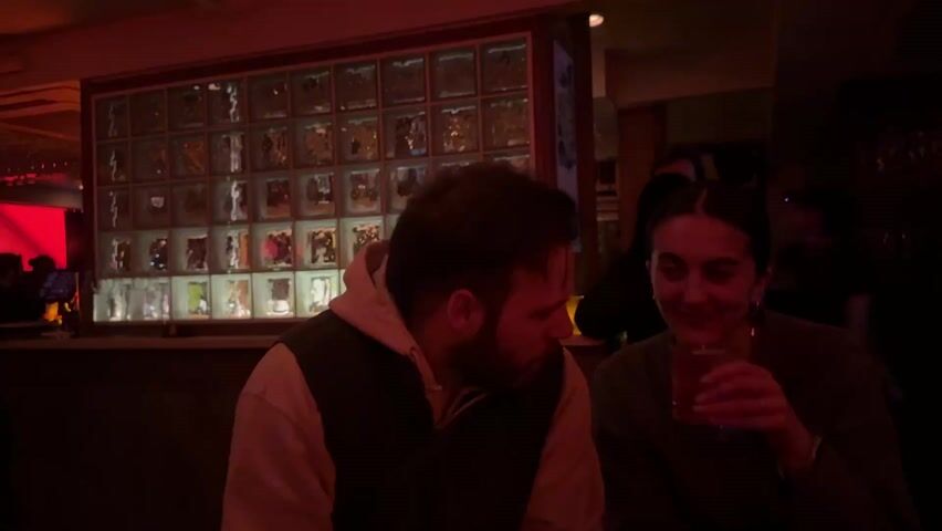 A couple is sitting at a bar. The man in a hoodie vest puts his hands on his face. The woman in the green sweater is smiling and drinking a reddish-brown beverage. There is a wall of lighted glass cubes behind the bar. A bartender walks behind them to the left. Other bar patrons are behind them as well. The lighting is dim and reddish.