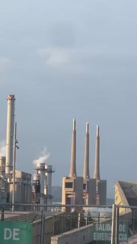 The video shows a view of an industrial area under a cloudy sky. There are several tall chimneys, including three that stand close together in a row. Smoke or steam is visible rising from one of the chimneys on the left. Various industrial buildings are visible, along with fencing and signs in the foreground. The overall tone is neutral, depicting an industrial landscape on what appears to be a typical day.