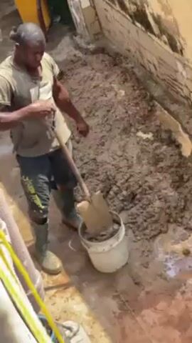 A man wearing a t-shirt, shorts, and rubber boots is using a shovel to mix mud in an outdoor area. He is standing in the mud, which covers a rectangular space. A white bucket sits next to him, partially filled with the mud. A yellow container is visible in the background, and the area is bordered by a wall.