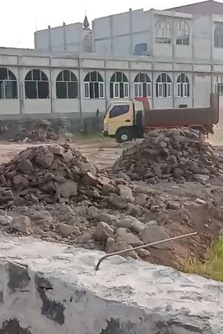 A yellow dump truck with a red bed is visible on a construction site. The truck is moving from left to right across the frame. There are large piles of rocks and dirt scattered around the site. In the background, a building with arched windows is under construction. The sky is overcast. The video is shaky and appears to be filmed from a handheld device.