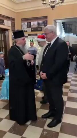 In an indoor setting with a checkered floor, a cleric wearing a black robe and headdress is engaged in conversation with a man in a black suit. A young girl in a blue princess dress stands to the left of the cleric holding a decorated box. Several other people are visible in the background, including one wearing a Santa hat. The room is decorated with a chandelier and wall posters. The cleric gestures with his hand as he speaks to the man in the suit, who listens attentively. The atmosphere appears to be calm and orderly, possibly during a community or religious event.