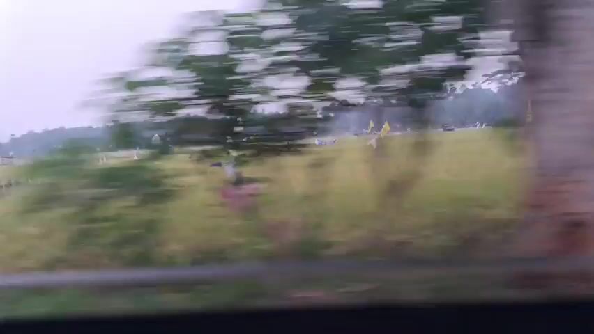 The video shows a rural landscape with a field of rice paddies dotted with scarecrows and flags. Buildings and trees are visible in the background. The camera is moving, suggesting it is taken from a vehicle. The sky is overcast and the overall lighting is dim, indicating it might be early morning or late afternoon. The video progresses to show more developed areas with shops and motorcycles parked outside. One shop has a prominent red and white sign that reads "J&T Express". The video ends by showing more buildings with shops and motorcycles, with some tables and chairs outside one establishment.