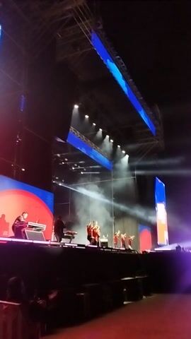 The video captures a dynamic night-time concert or live performance, likely taking place outdoors in Maipú, Chile. The stage features an elaborate setup, characterized by robust metal scaffolding and several large LED screens. Two prominent, elongated screens are positioned diagonally on the left, displaying vivid blue and white patterns. On the right side of the stage, additional screens are visible, including one at the lower right that shows a close-up of a performer, possibly a DJ or keyboardist, dressed in dark clothing.

The stage is brightly lit by numerous spotlights and other stage lights, casting intense white and red beams that cut through the smoky atmosphere. Plumes of white smoke or fog frequently billow across the stage, enhancing the visual effects of the lighting. Multiple performers are distributed across the stage; while most are somewhat obscured by the lighting and smoke, their presence indicates a full band setup with instruments like drums and keyboards. A large red structure on the far right partially frames the scene. The overall ambiance is energetic and immersive, typical of a large-scale musical event. Although the audience is not visible, their presence is implied by the setup. There is no clearly readable text visible on any of the screens or stage elements.