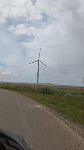 A car travels along a narrow asphalt road. To the right of the road, a field of tall green grass stretches to the horizon, dotted with numerous wind turbines. The turbines have three blades and are white in color. Overhead, a cloudy sky blankets the scene. An electric pole is visible in the background, and a car dashboard is briefly visible in the bottom left corner of the frame.