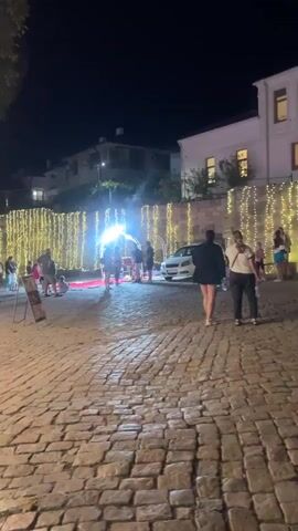 The video shows a bustling night scene in Sozopol, Bulgaria. The setting appears to be a charming cobblestone street lined with buildings and trees. Numerous strings of warm, golden lights are draped across the scene, illuminating a lively atmosphere.

Several people are visible, mingling and enjoying the evening. In the foreground, two women are walking away from the camera on the cobblestone street. Further into the scene, a group of people are gathered near a brightly lit archway adorned with more lights. Some individuals are standing and chatting, while others appear to be walking through the illuminated structure. To the left, a few children are playing or standing near some decorative elements.

Various objects are present, including a white car parked on the right side of the street. On the left, a motorcycle is parked, and a wooden sign on a stand is visible. Towards the right and center, there are market stalls or food vendors. One stall, partially visible, displays an array of food items under a tent, with bright lights illuminating the products. Another stall further back appears to be selling baked goods or similar items, with stacks of bread-like products on display.

The time of day is clearly nighttime, indicated by the darkness and the prominent use of artificial lighting. The weather appears to be pleasant, as people are dressed in casual clothing and the outdoor activity suggests comfortable conditions. The overall emotion conveyed is one of festivity and enjoyment, with the bright lights and gathering of people suggesting a special event or a lively evening in the town. No discernible text is visible in the video.