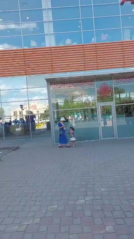The video shows the exterior of a modern shopping mall with a glass facade and orange accents. A woman in a blue dress and white hat stands near the entrance with a young child. Several people are entering and exiting the mall through automatic doors. The ground is paved with interlocking stones. Beyond the mall, a parking lot is visible with several cars. The sky is blue with scattered white clouds. A woman in a headscarf and brown skirt stands facing away from the camera, holding a yellow shopping bag. The video pans to the right, revealing more of the parking lot and surrounding area.