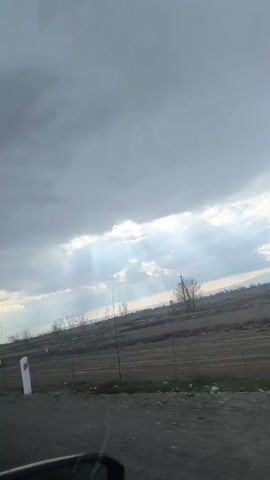 The view from inside a moving vehicle shows an overcast, gray sky with beams of sunlight breaking through. The landscape consists of a field with sparse vegetation and what appears to be tilled earth. Power lines and utility poles are visible across the field. The road is gravelly, and the side mirror of the vehicle is partly visible in the foreground.
