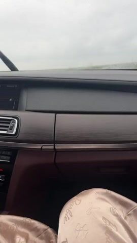 The video captures a first-person perspective from inside a car driving on a highway in Iabalkovo, Bulgaria. The weather is overcast and rainy, with visible raindrops on the windshield. The car is on a multi-lane road flanked by green fields and guardrails, with distant structures (possibly power lines) visible under a cloudy sky. The car's dashboard is prominent, featuring dark and wood-textured panels. A car display shows "THEVOICE TP." In the bottom right corner, a light-colored fabric item with the word "Barbie" printed on it is partially visible. Music is playing throughout the recording.