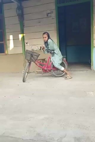 A young girl in a hijab and a checkered skirt is sitting on a pink bicycle. She is holding onto the handlebars, with her feet on the ground. The bicycle is leaning to the right. The girl is looking at the camera. The background is a wooden house with a door. The ground is concrete.