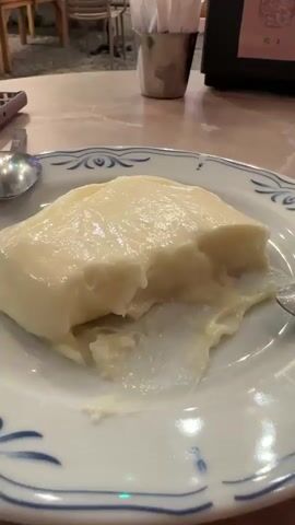 The video opens with a close-up of a white plate with a serving of what appears to be a creamy dessert, possibly pudding or mousse, with a bite taken out of it. A spoon is visible to the left of the plate, and another spoon is partially visible to the right. The plate has a blue and white decorative rim. The background is blurred but shows an outdoor dining setting. As the video progresses, the camera pans slightly to the left, revealing more of the environment. Tables and chairs are visible, suggesting a cafe or restaurant. In the background, a building facade is visible, decorated with string lights in blue, green, and white. A person wearing a blue shirt and dark shorts is standing inside a doorway or counter area, appearing to interact with something or someone. Another person is seated at a table to the right, partially obscured. The ground is paved with cobblestones. A black box, possibly a napkin dispenser or payment terminal, is on a table in the foreground. An ashtray is next to the black box. The overall scene depicts a casual dining environment at dusk or in the evening, with ambient lighting from the string lights.