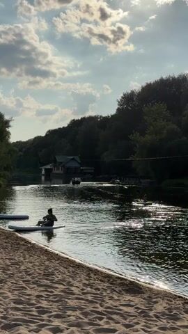 A person sits on a paddleboard in the water near a sandy beach. The person is wearing a dark shirt and shorts. The paddleboard is white. Two other paddleboards are partially visible on the sand to the left of the person. The water is a river or lake. In the background, there is a house or building on the water, and trees line the opposite bank of the water. The sky is cloudy with sunlight breaking through. The scene appears to be a natural outdoor setting.