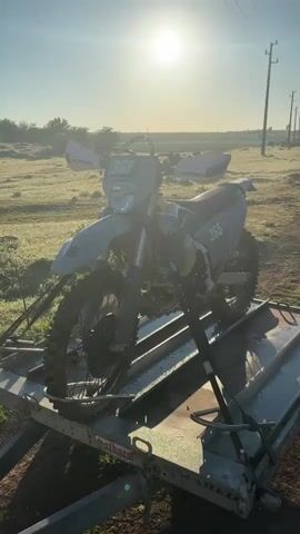 A motorcycle is securely strapped to a metal trailer against the backdrop of a wide open field under a bright, hazy sunlight. The motorcycle is predominantly gray and features the number 365 prominently displayed on the front and side. The tires show signs of recent off-road use, covered in mud. The trailer is parked on a grassy area that seems to be covered in morning dew. Power lines stretch across the landscape in the distance. The light is strong, creating lens flare and partially obscuring some details of the scene.