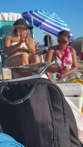The video shows a beach scene with two women in the background. The woman on the left is wearing a white bikini and a baseball cap. She is sitting in a beach chair and appears to be clapping. The woman on the right is wearing a floral print dress and sunglasses. She is also sitting and clapping, with her hands together in front of her. Both women are looking towards the right side of the frame. In the foreground, a black backpack is partially visible, with a blue object to its left. Beach umbrellas are visible in the background, along with other beachgoers and the ocean. The lighting suggests it is a sunny day.