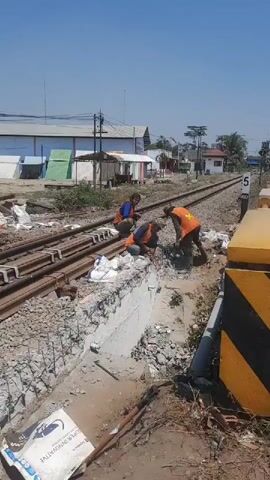 Three workers in orange safety vests are repairing railroad tracks. The worker on the left is sitting and appears to be supervising. The middle worker is kneeling and is using a tool to manipulate rebar. The worker on the right is standing and is using a jackhammer to break up concrete. The area is littered with debris, including broken concrete and bags of material. There are railroad tracks in the background, as well as some buildings and trees. The sky is blue and it appears to be a sunny day.