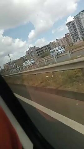 The video, taken from a moving vehicle, shows a multi-lane highway with various buildings and infrastructure alongside it. The sky is partly cloudy with a blue sky visible in patches. Several buildings, including apartment complexes and buildings under construction, are visible in the background. On the highway, there are vehicles of various sizes, including vans and cars, traveling in both directions. The edge of the highway features a metal guardrail and patches of brown grass and vegetation.  The perspective is from the passenger side of the vehicle, giving a side view of the highway and the surrounding landscape.
